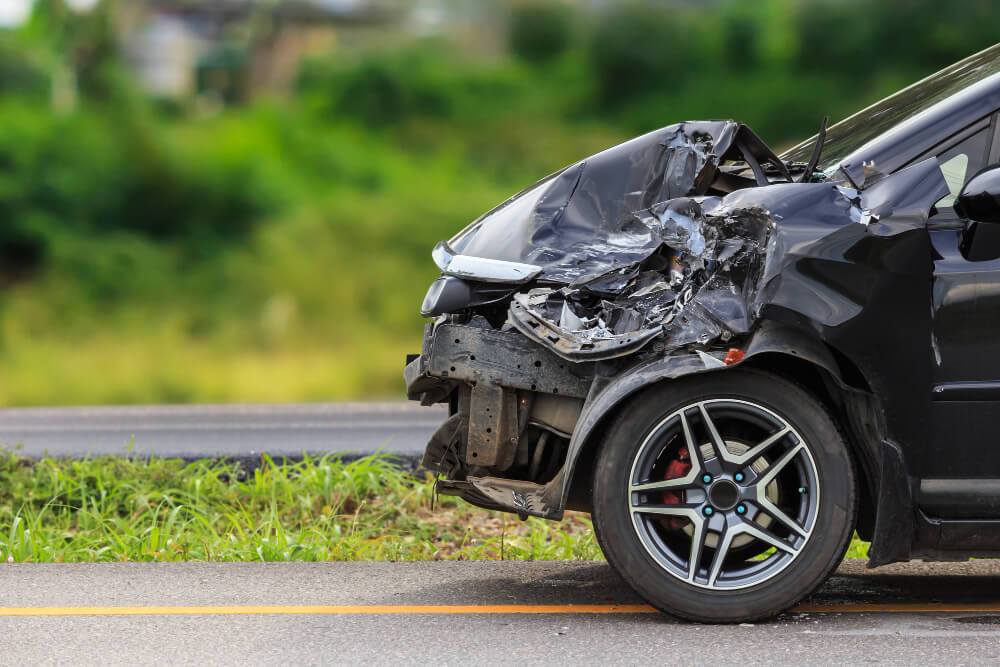 Ein schwarzes Auto mit erheblichen Frontschäden steht am Straßenrand vor Grünflächen und eignet sich für ein Schadengutachten