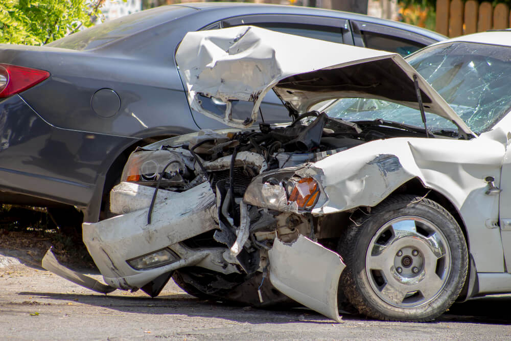 Ein weißes Auto mit schweren Frontschäden nach einem Unfall steht neben einem unbeschädigten grauen Auto am Straßenrand