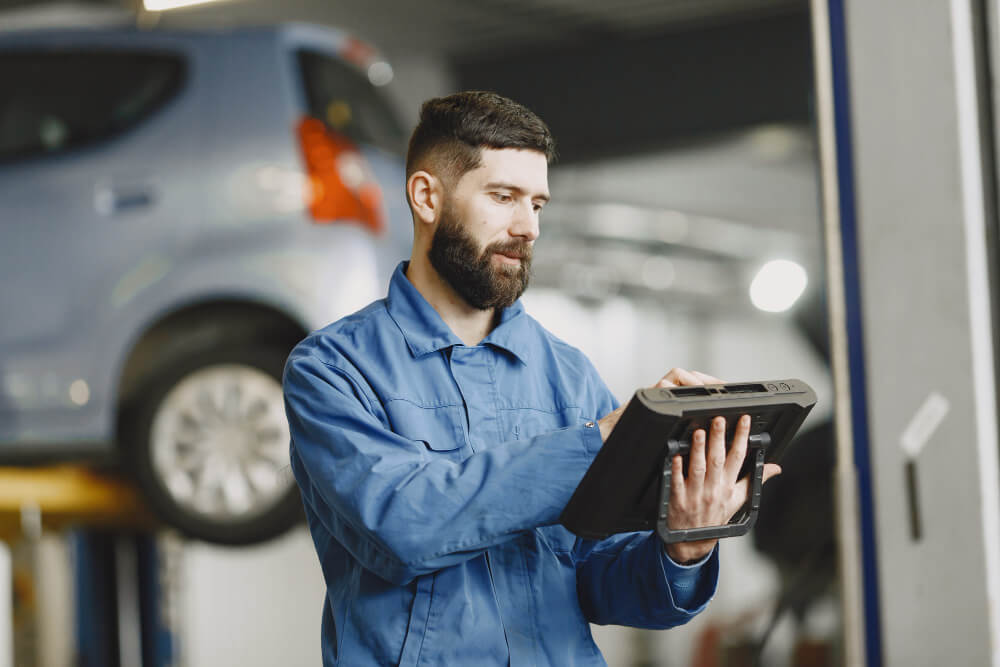 Ein Mechaniker im blauen Overall nutzt ein Tablet für ein Unfallgutachten, während im Hintergrund ein Auto in der Werkstatt angehoben wird.