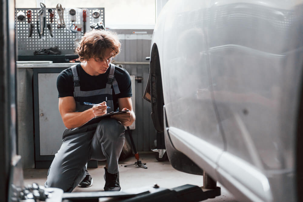 Ein Mechaniker kniet neben einem Auto und schreibt in einer Werkstatt mit an der Wand hängendem Werkzeug auf ein Klemmbrett