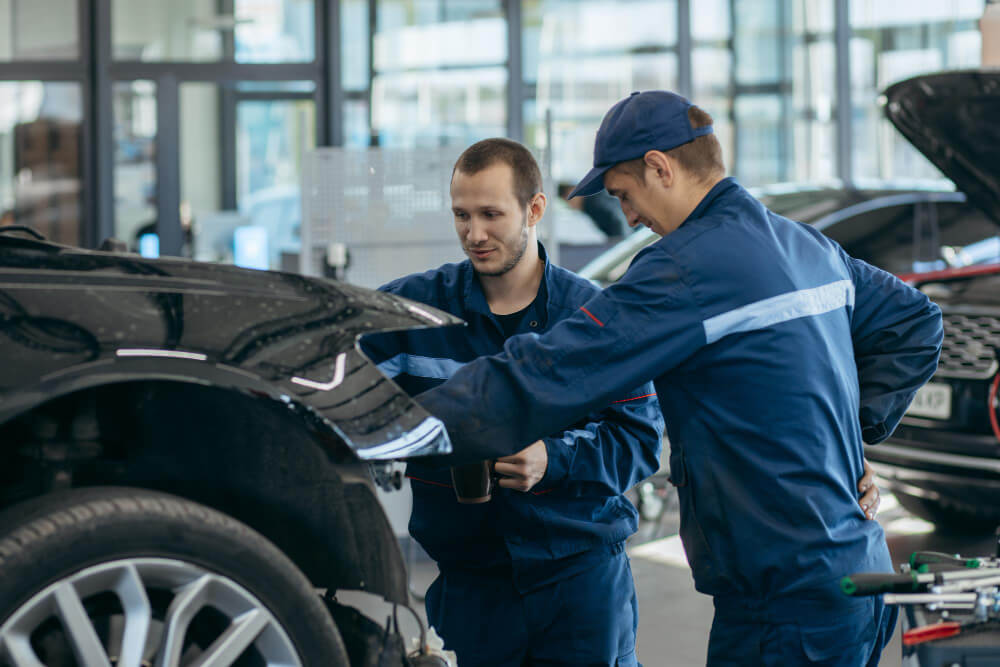 Zwei Mechaniker in blauer Arbeitskleidung prüfen den Motor eines schwarzen Autos in einer hellen Autowerkstatt