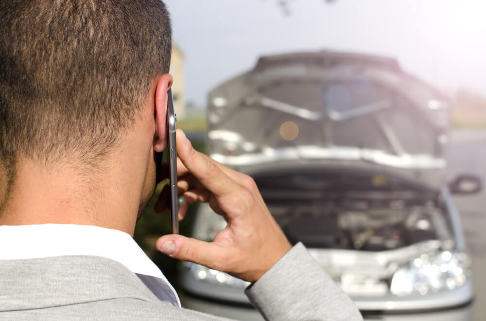 Ein Mann telefoniert mit seinem Handy, im Hintergrund steht ein beschädigtes Auto, vermutlich benötigt er ein Unfallgutachten